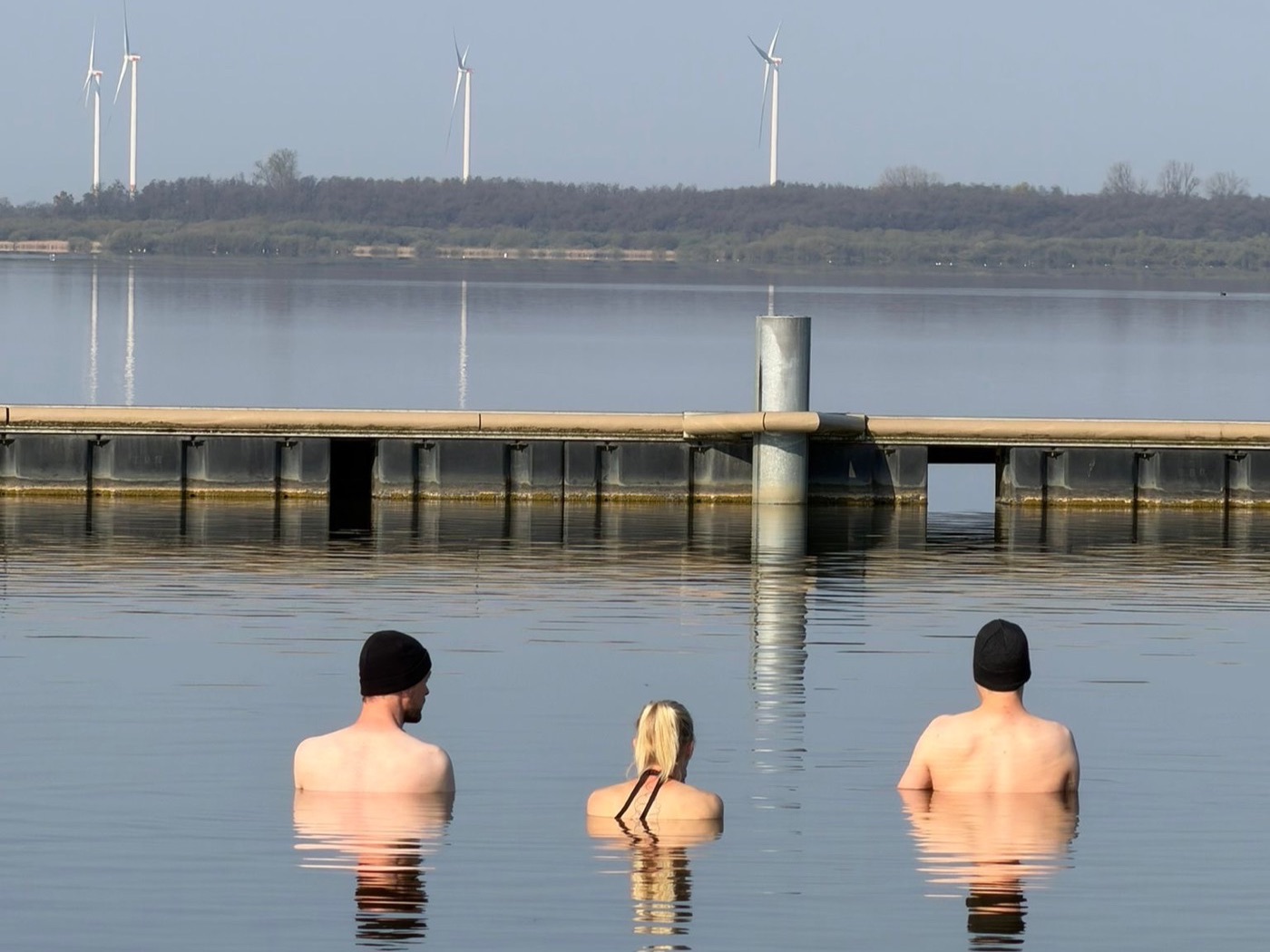 Three cold-water swimmers in Lake Dümmer, jetty and wind turbines behind